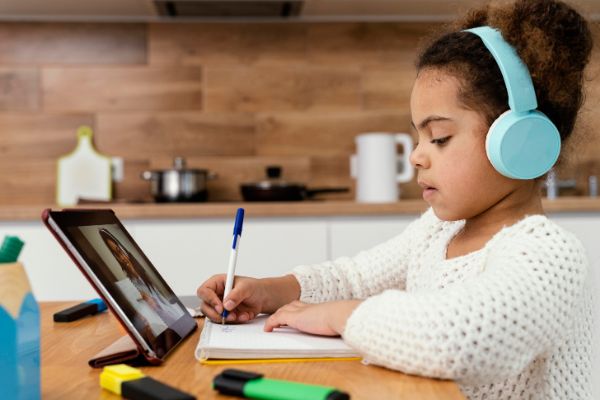 Criança na mesa olhando para a tela de um computador escrevendo no caderno